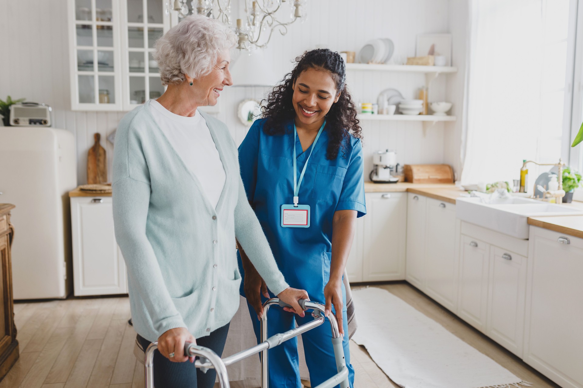 African american medical nurse volunteer helping senior female at home, teaching her to walk with walker, smiling and giving instructions how to use device, standing together in cozy kitchen